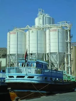 Drei Zementsilos auf Stahlgerüst am Hafen, davor ein traditionelles Holzschiff